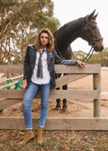 A woman wearing a black leather jacket, white shirt, gray tie, blue jeans, and tan boots leans against a wooden fence next to a dark brown horse in an outdoor paddock.