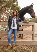 A woman wearing a black leather jacket, white shirt, gray tie, blue jeans, and tan boots leans against a wooden fence next to a dark brown horse in an outdoor paddock.