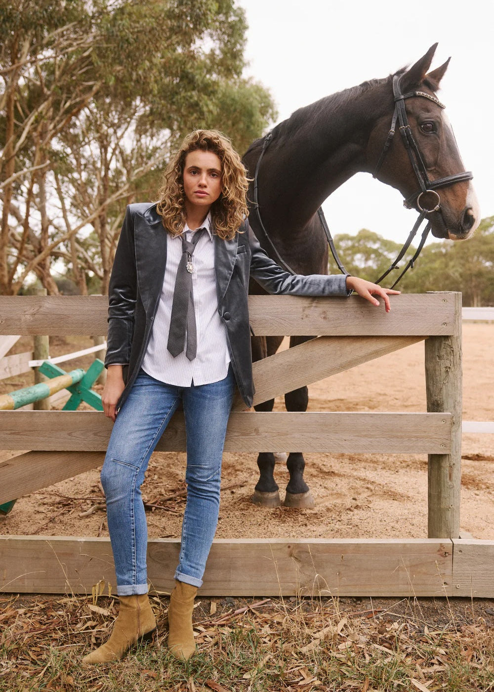 A woman wearing a black leather jacket, white shirt, gray tie, blue jeans, and tan boots leans against a wooden fence next to a dark brown horse in an outdoor paddock.